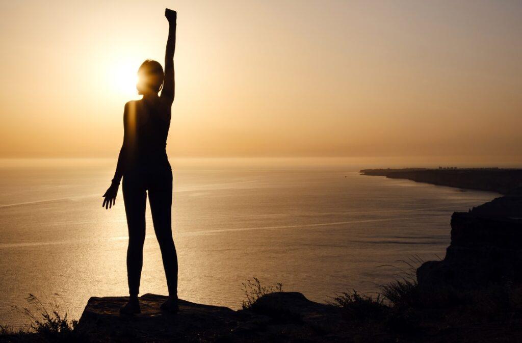 Femme debout face à la mer au coucher de soleil, levant le bras en signe de victoire